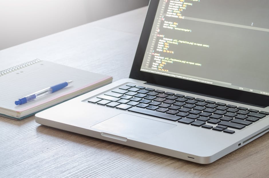 A close-up view of a silver laptop with a black keyboard displaying lines of code on a dark screen, placed on a wooden desk. To the left of the laptop, there is an open notepad with a blue and white p