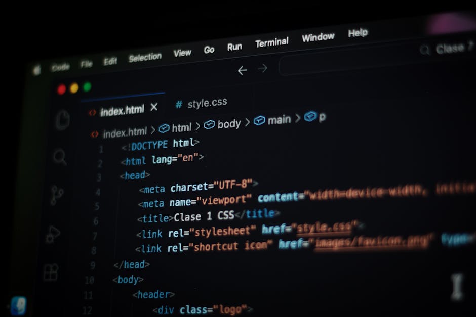 A close-up view of a silver laptop with a black keyboard displaying lines of code on a dark screen, placed on a wooden desk. To the left of the laptop, there is an open notepad with a blue and white p